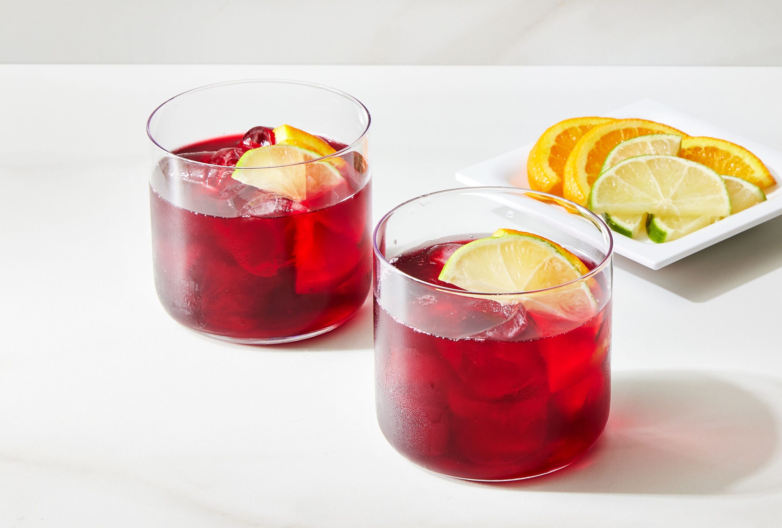 Two glasses of hibiscus tea with slices of lemon floating on top sit on a white countertop.