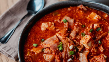 A close-up on ZCounter's kimchi soup, served inside a black bowl next to a bowl of rice and a spoon and napkin.