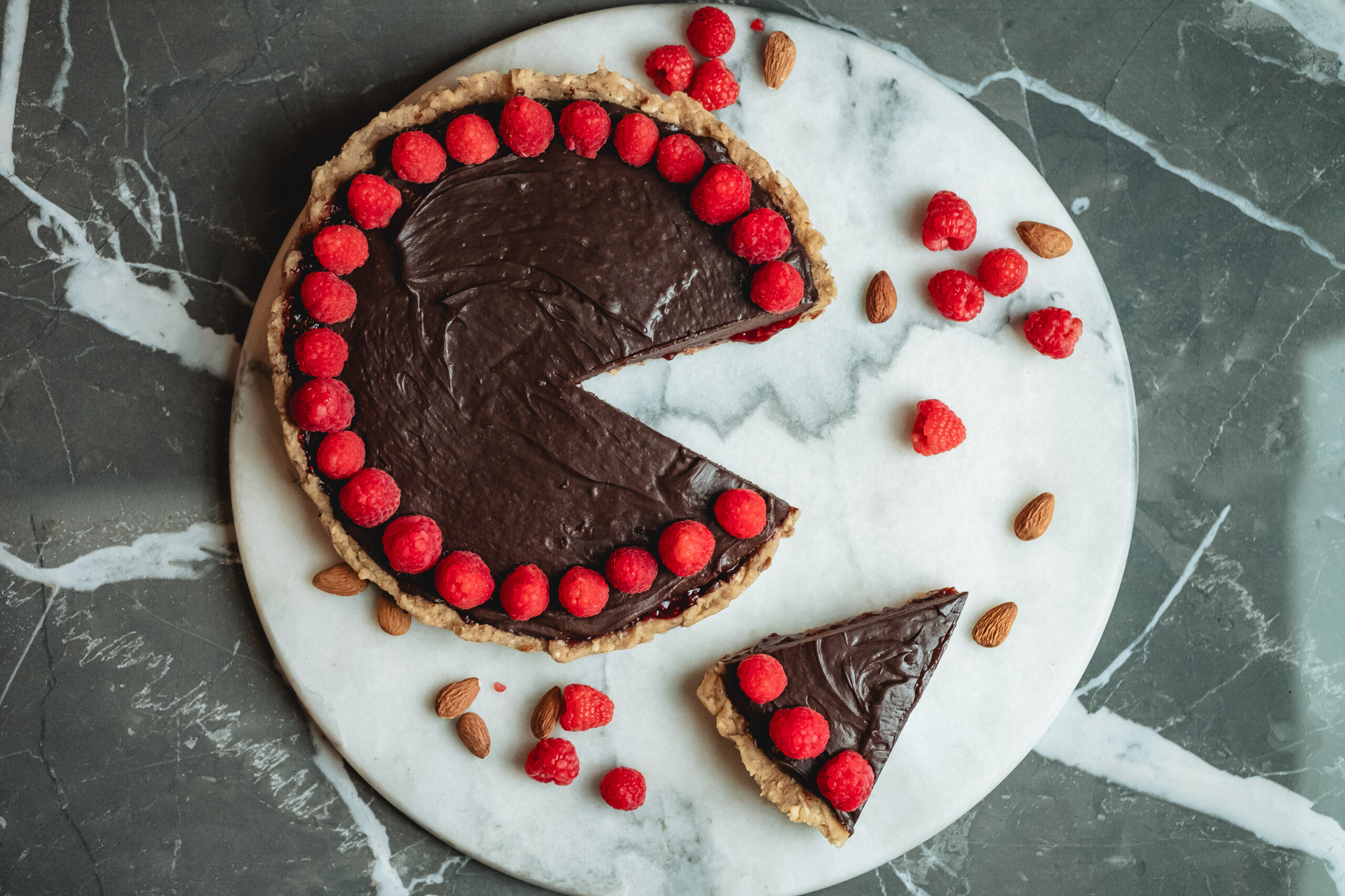 An overhead view of a chocolate raspberry tart sitting on a white marvel plate set on top of a black marble countertop, decorated with chocolate frosting and raspberries lining the perimeter of the circle cake.