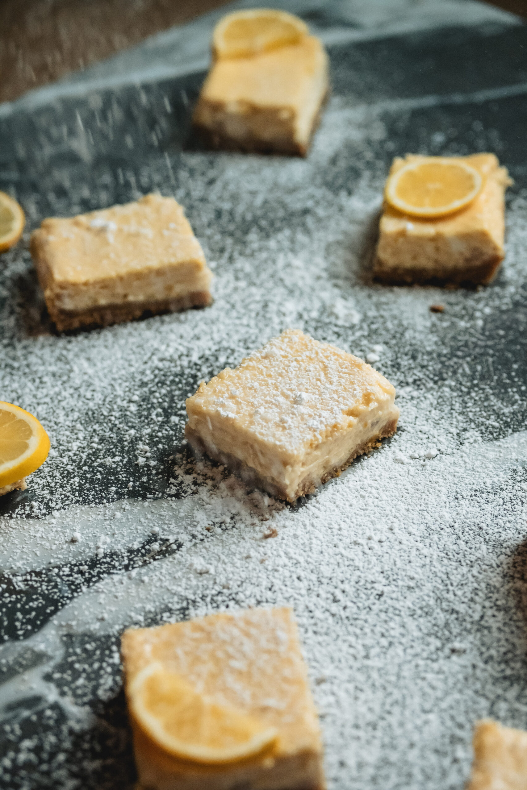 A side view of lemon bars and lemon slices sit scattered across a marble counter top sprinkled with powdered sugar.