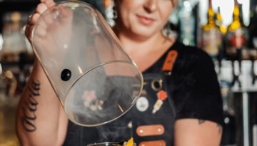 A female bartender unveils a dark craft cocktail in a small air of smoke inside Fork & Vines.