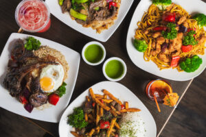 An overhead view of four specialty dishes from Kapi Honda, sitting on a brown table with two green sauces and two drinks set in the middle.